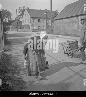 Unbefristetes Foto einer barfüßigen alten Buckelfrau in Schürze und Kapuze, während sie Milch in Oberlußland, ehemaliger sowjetischer Besatzungszone (SBZ, heute Freistaat Sachsen) sammelte. Die Frau hält ein kleines Milchkännchen in der rechten Hand. Rechts, ein Mann in Arbeitskleidung, Clogs und einer flachen Mütze, der einen Wagen mit Korb schiebt. Im Hintergrund die Dorfstraße und ein Gasthaus. Stockfoto