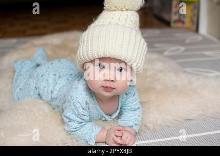 Porträt eines Babys in einem Winterhut, das auf einem Schafvorleger krabbelt. Süßes weißes Baby mit dunklen Augen Porträt Stockfoto