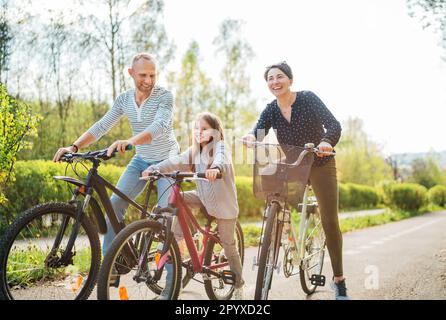 Lächelnder Vater und lächelnde Mutter mit Tochter während der Sommerfahrradrundfahrt. Sie genießen die Zusammengehörigkeit im Sommer-Stadtpark. Glückliche Elternschaft und ch Stockfoto