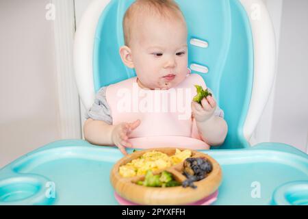 Baby isst Brokkoli. Säuglingsprothetik. Entwöhnung. Gesunde Ernährung. Kaukasisches Mädchen, das in einem Hochstuhl sitzt und ihr Mittagessen isst Stockfoto