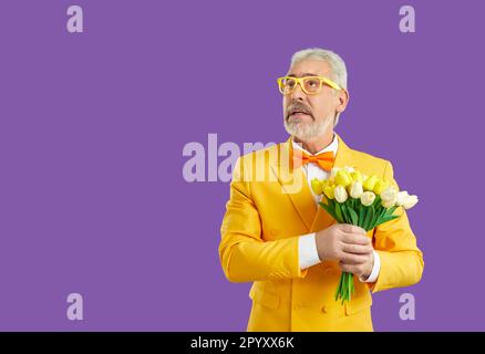 Lustiger, alter, grauhaariger Mann mit einem Strauß Tulpen in den Händen sieht in lila Studio auf Stände Stockfoto