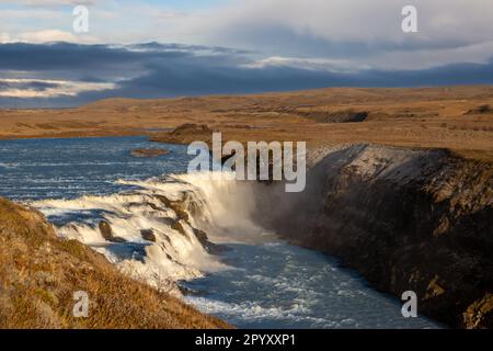 Berühmter Teil des goldenen Kreises. Wasserfall Gullfoss, umgeben von Klippen. Goldene Farbe der Natur am frühen Abend. Wolkiger Himmel Stockfoto