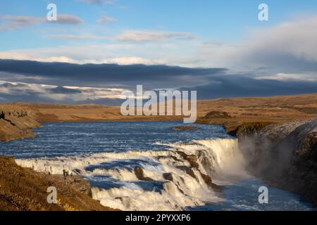 Berühmter Teil des goldenen Kreises. Wasserfall Gullfoss, umgeben von Klippen. Goldene Farbe der Natur am frühen Abend. Wolkiger Himmel Stockfoto