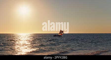 Sonnenuntergang über dem ruhigen Meer, kleines Segelpiroga-Boot mit Silhouette eines unbekannten Mannes in der Ferne Stockfoto