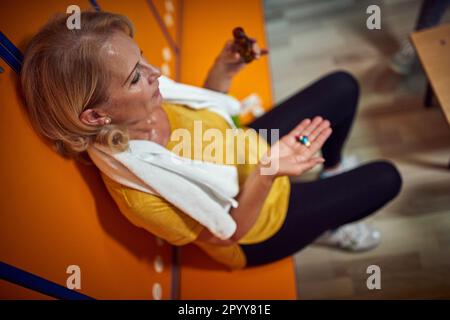 Horizontale Top-Aufnahme einer Seniorin im Umkleideraum Holdigpillen, die Medikamente und Vitamine nach dem Training nehmen. Das Seniorenlebenskonzept. Stockfoto