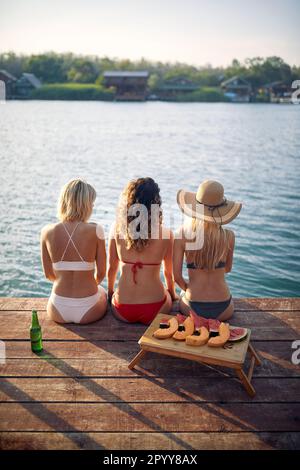 Drei junge Frauen in Badeanzügen, die auf einem Holzsteg sitzen und sich unterhalten und den Sommertag genießen. Rückansicht. Urlaub, Zusammengehörigkeitskonzept. Stockfoto