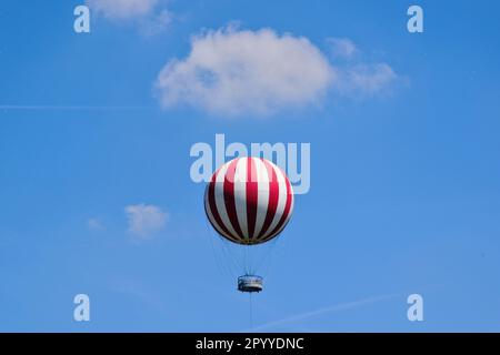 Isolierter rot-weißer Abenteuerballon hoch oben. Hängekorb und Aussichtsplattform mit Menschen. Aufhängungsseile. Blauer Himmel, Wolken. Besichtigungstour. Stockfoto