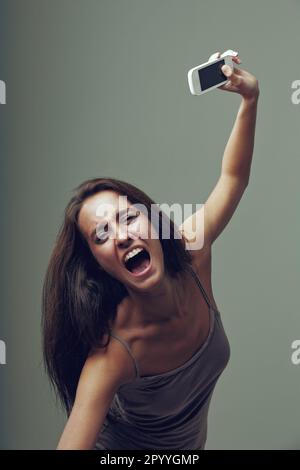 Wütendes Mädchen wirft ihr Handy vor Wut. Emotionales Porträt einer jungen Frau isoliert auf neutralem Hintergrund. Sie denkt: "Ich habe diese sma satt Stockfoto