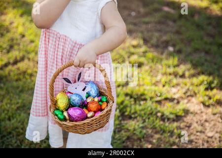 Ein kleines Mädchen zu Ostern mit einem Korb mit in Folie verpackten Ostereiern, die im Park gefunden wurden Stockfoto