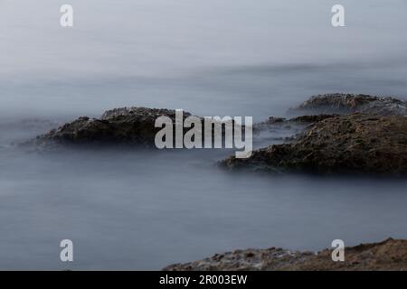 Eine malerische Strandszene mit einem Vordergrund aus großen Felsen mit Regenwasser, das über sie und ins Meer dahinter dringt Stockfoto