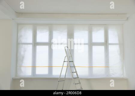 Fenster mit Plastikfolie und Leiter im renovierten Zimmer Stockfoto