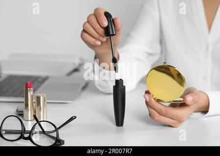 Junge Frau, die einen kosmetischen Taschenspiegel und Mascara an einem weißen Tisch im Haus benutzt, Nahaufnahme Stockfoto