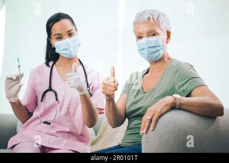 Mehr als ein Arzt...Porträt einer maskierten Ärztin und ihrer maskierten Patientin, die nach einer Injektion zu Hause die Daumen nach oben zeigen. Stockfoto