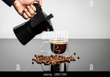 Eine Person, die frischen Kaffee aus einer Kanne in eine Tasse mit Kaffeebohnen gießt Stockfoto