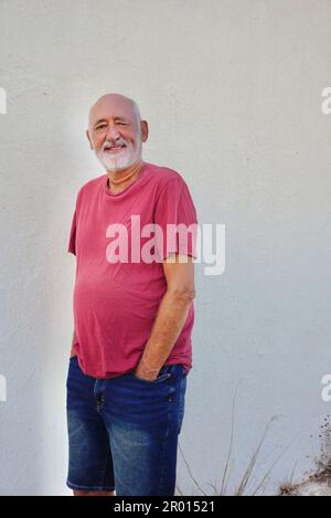 Ein glücklicher alter Mann, der ein rosa Hemd trägt und an der weißen Wand steht Stockfoto