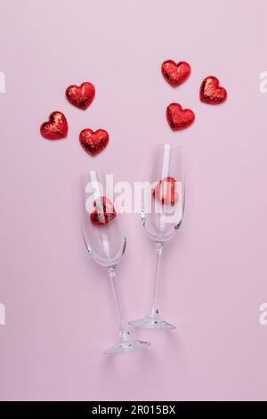 Romantische valentinstagskarte mit zwei klickenden Champagnergläsern und einem Spritzer roter herzförmiger Konfetti auf pinkfarbenem Hintergrund. Draufsicht. Fla Stockfoto