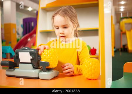 Kind im Kindergarten, im Spielzimmer, mit Spielzeug spielen, lernen. Spiele im Geschäft, Gemüse und Obst, Kasse und Waage. Wiegt Ananas. Stockfoto