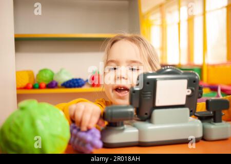 Kind im Kindergarten, im Spielzimmer, mit Spielzeug spielen, lernen. Spiele im Geschäft, Gemüse und Obst, Kasse und Waage. Komisch Stockfoto