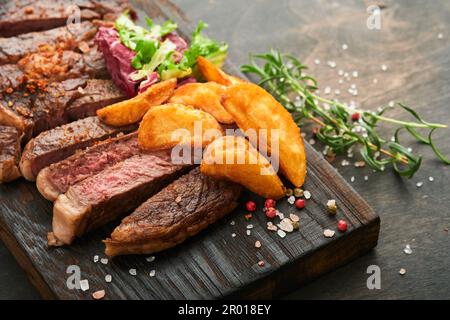 Steaks. Geschnittenes gegrilltes Fleischsteak New York, Ribeye oder Chuck Roll mit garniertem Salat und pommes frites auf schwarzem Marmorbrett auf altem Holzbrett Stockfoto