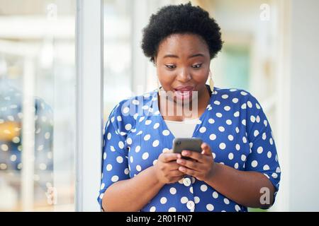 Du hast hart gearbeitet, jetzt siehst du zu, wie Millionen reinkommen. Eine junge Geschäftsfrau, die schockiert aussah, als sie ein Smartphone in einem modernen Büro benutzte. Stockfoto