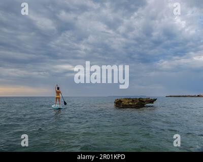 Frau paddelt auf einem Surfbrett unter einem dramatischen Himmel, Estalella, Küste von Llucmajor, Mallorca, Balearen, Spanien. Stockfoto