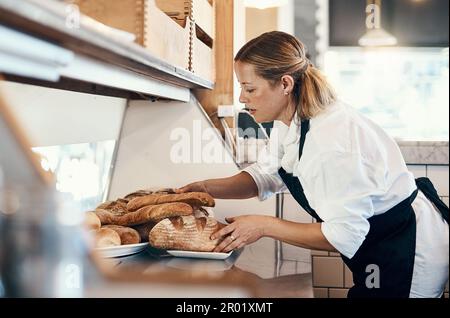 Ihr Brot steigt weit über den Rest. Eine reife Frau, die eine Auswahl an frisch gebackenem Brot in ihrer Bäckerei ausstellt. Stockfoto