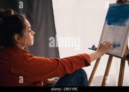 Attraktive, stilvolle junge Frau, inspiriert von einem Look, der Pinsel und Farbmalerei auf Leinwand hält, wobei Ölfarbe vor der Staffelei steht Stockfoto