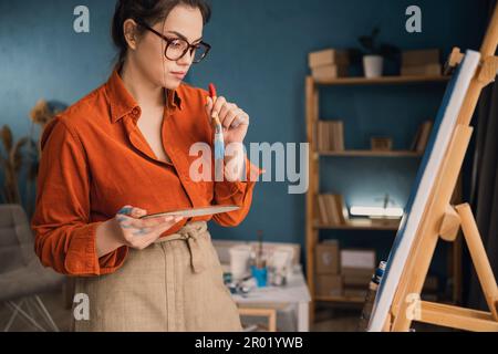 Seitenansicht fokussierte ethnische Frau in lässiger Kleidung steht neben der Leinwand hält eine Palette mit Farben in ihren Händen schaut auf die Leinwand und denkt darüber nach Stockfoto