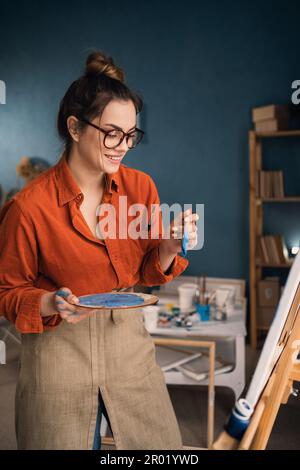 Seitenansicht fokussierte ethnische Frau in lässiger Kleidung steht neben der Leinwand hält eine Palette mit Farben in ihren Händen schaut auf die Leinwand und denkt darüber nach Stockfoto