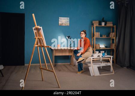 Inspirierende, konzentrierte Latinofrau in der Schürze zeichnet Bilder mit Farben im modernen Studio oder Wohnzimmer. Eine nachdenkliche Künstlerin sitzt und schaut sich das an Stockfoto