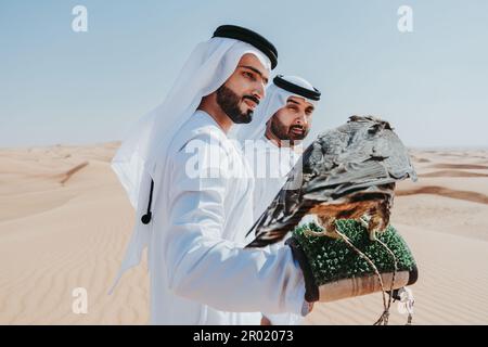 Zwei Männer aus dem Mittleren Osten, die traditionelle arabische kandura tragen, sich in der Wüste verbinden und einen Falken halten, arabische muslimische Freunde, die sich dort treffen Stockfoto