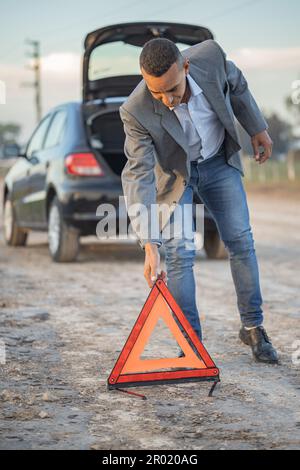 Ein junger Latino-Mann, der ein Warndreieck hinter einem kaputten Auto platziert. Stockfoto