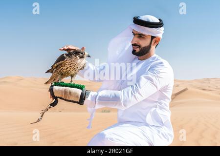 Ein Mann aus dem Mittleren Osten, der in der Wüste traditionelle arabische kandura trägt und einen Falken hält - arabisch-muslimischer Erwachsener in den Sanddünen Stockfoto