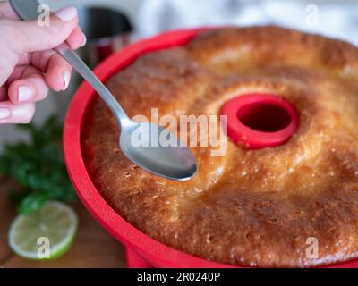 Frisch gebackenes Savarin in rotem Silikonkuchenform wird mit Sirup gegossen, selektiver Fokus. Rum-getränkter, kreisförmiger Kuchen, Nahaufnahme Stockfoto
