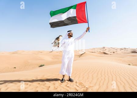 Ein Mann aus dem Nahen Osten, der in der Wüste traditionelle arabische kandura aus den emiraten trägt und einen Falken und die Flagge der Vereinigten Arabischen Emirate in der Hand hält - eine Erwachsene arabische muslimische Person Stockfoto