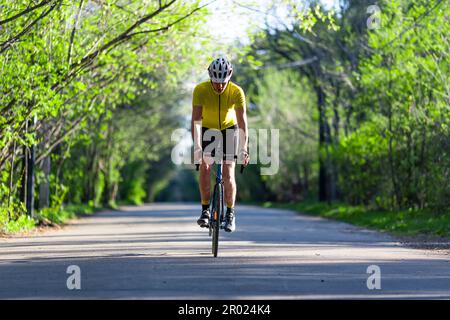 Ein junger Mann mit Schutzhelm und Aktivkleidung, der im Sommer auf einer befestigten Straße im grünen Wald fährt. Regelmäßiges Training und gesunder Lebensstil. Stockfoto