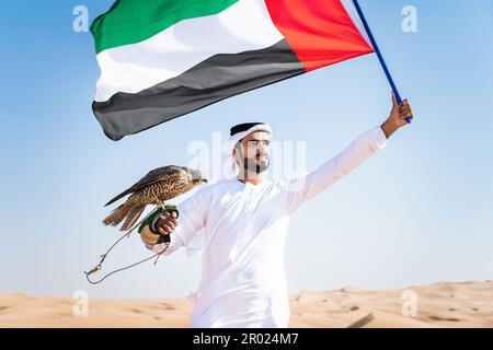 Ein Mann aus dem Nahen Osten, der in der Wüste traditionelle arabische kandura aus den emiraten trägt und einen Falken und die Flagge der Vereinigten Arabischen Emirate in der Hand hält - eine Erwachsene arabische muslimische Person Stockfoto