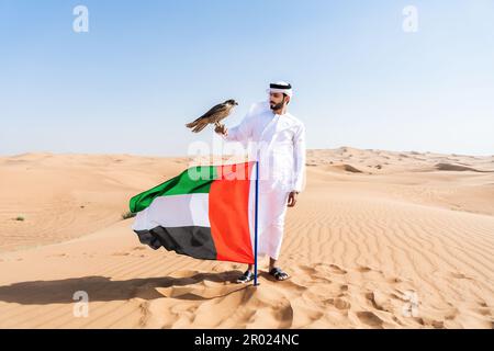 Ein Mann aus dem Nahen Osten, der in der Wüste traditionelle arabische kandura aus den emiraten trägt und einen Falken und die Flagge der Vereinigten Arabischen Emirate in der Hand hält - eine Erwachsene arabische muslimische Person Stockfoto