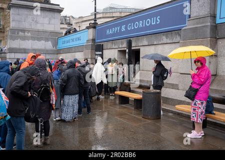 Unter dem Banner, das „glücklich und glorreich“ verkündet, stehen Mitglieder der öffentlichen Schlange für die Toiletten, nachdem sie die Krönungszeit von König Karl III. Am Trafalgar Square am 6. Mai 2023 in London, England, angeschaut haben. Stockfoto