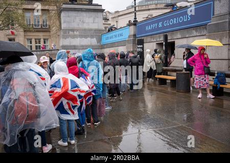 Unter dem Banner, das „glücklich und glorreich“ verkündet, stehen Mitglieder der öffentlichen Schlange für die Toiletten, nachdem sie die Krönungszeit von König Karl III. Am Trafalgar Square am 6. Mai 2023 in London, England, angeschaut haben. Stockfoto