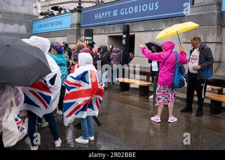 Unter dem Banner, das „glücklich und glorreich“ verkündet, stehen Mitglieder der öffentlichen Schlange für die Toiletten, nachdem sie die Krönungszeit von König Karl III. Am Trafalgar Square am 6. Mai 2023 in London, England, angeschaut haben. Stockfoto