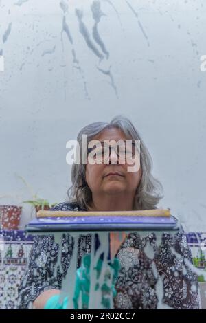 Nahaufnahme einer weißhaarigen Frau, die seifenbedeckte Fenster mit einem Abzieher reinigte Stockfoto