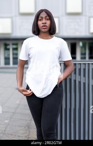 Mühelos cool: Ein Porträt einer stilvollen jungen schwarzen Frau mit grauem Gebäude als Hintergrund, die ein weißes T-Shirt und eine schwarze Hose trägt Stockfoto