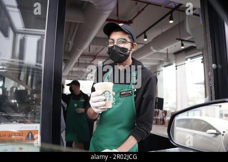 Starbucks Mitarbeiter geben Befehle mit fröhlichem Gesichtsausdruck und tragen eine Gesichtsmaske am Drive-in. Eisgekühlter weißer Mokka-Kaffee. Stockfoto