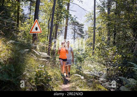 Mann und Frau wandern in infizierten Zeckenwäldern mit Warnschild. Risiko von Zecken-Borreliose und lyme-Borreliose. Stockfoto