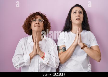 Hispanische Mutter und Tochter tragen ein legeres weißes T-Shirt auf pinkfarbenem Hintergrund und betteln mit den Händen zusammen mit dem Ausdruck der Hoffnung im Gesicht Stockfoto