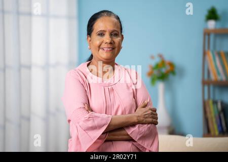 Porträtaufnahme einer glücklichen lächelnden, selbstbewussten indischen Seniorin, die mit verschränkten Armen zu Hause steht und die Kamera anschaut - Konzept der Zufriedenheit Stockfoto