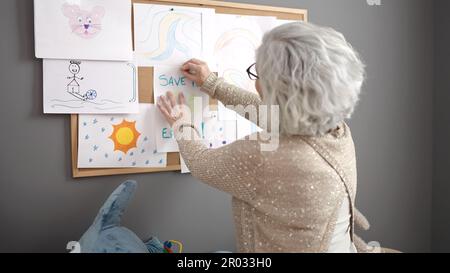 Eine Frau mittleren Alters mit grauhaariger Vorschullehrerin, die im Kindergarten eine Zeichnung auf Korkbrett hängt Stockfoto