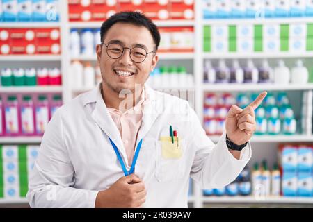 Chinesischer junger Mann, der in einer Apotheke arbeitet, mit einem großen Lächeln im Gesicht, der mit dem Finger zur Seite zeigt und in die Kamera schaut. Stockfoto