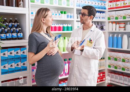 Mann und Frau, Apotheker und schwangere Klientin, die in der Apotheke ein Smartphone benutzen Stockfoto